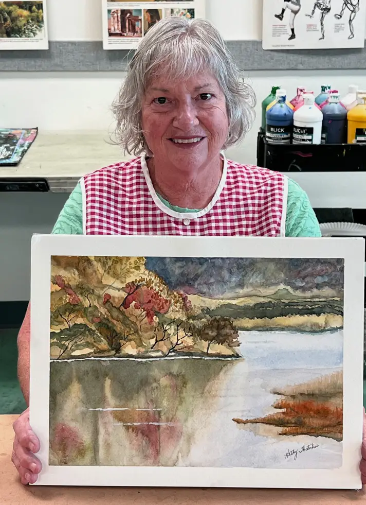 Elderly woman proudly showing her watercolor landscape of a lake and mountains during Art Class in Leander