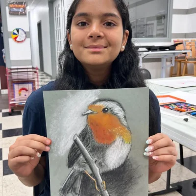 Young girl holding up a pastel drawing of a bird with black and white contrast and an orange chest during Art Class in South Austin