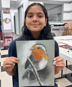 Young girl holding up a pastel drawing of a bird with black and white contrast and an orange chest during Art Class in South Austin