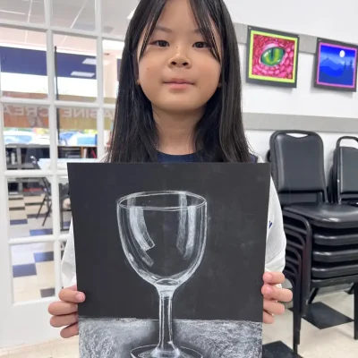 A young student proudly presents her artwork featuring a beautifully shaded pear set against a colorful abstract background during Art Class in Round Rock. The piece highlights her skill in value, contrast, and creative composition — blending realism with imaginative design in a supportive classroom setting.